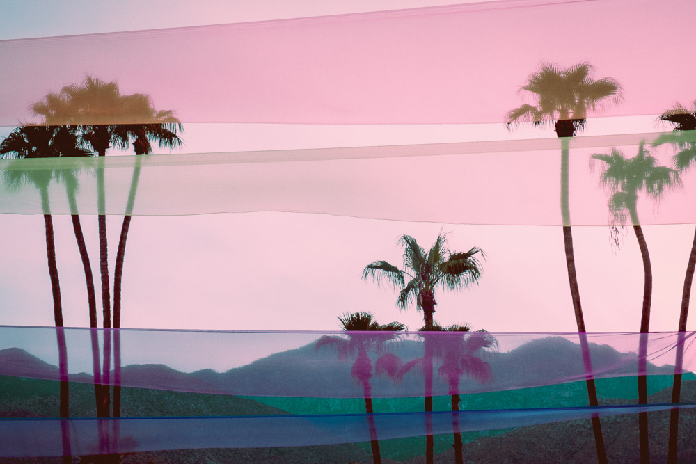 view of three sets of palms trees with the mountains in the back and coloured ribbons in the sky