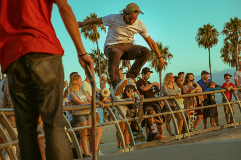 skater jumping in the air and other skater with red shirt watching him with people and palm trees