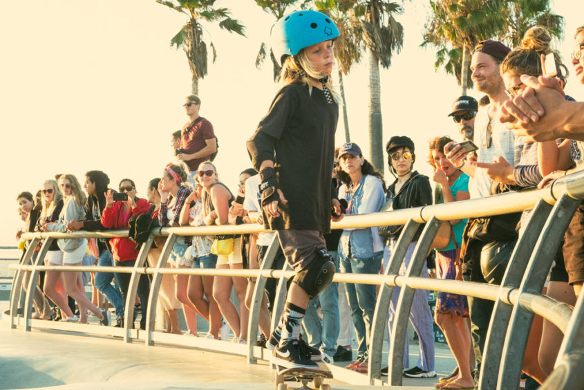 little blond girl with helmet at the venice beach skate park with people watching her
