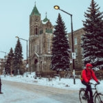 street with snow, church a person crossing and another with a red coat on a bike